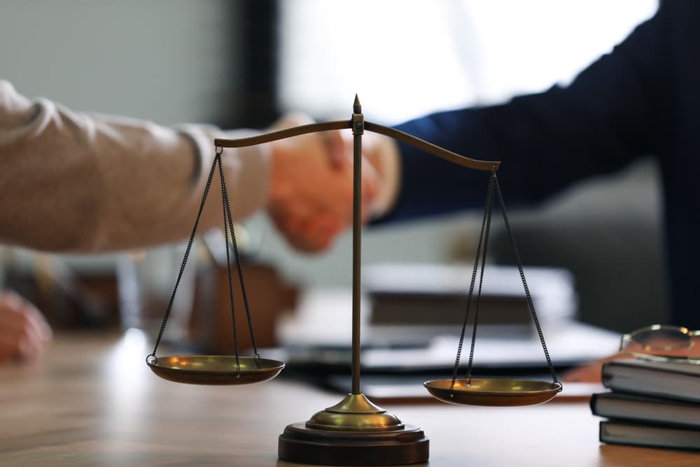 A male lawyer shaking hands with a client in an office, with the scales of justice in focus. A male lawyer shaking hands with a client in an office, with the scales of justice in focus.
