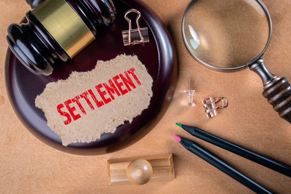 Legal settlement concept with judge’s gavel, magnifying glass, and settlement document on desk. Legal settlement concept with judge’s gavel, magnifying glass, and settlement document on desk.