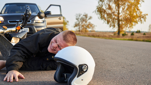 A man lies injured on the road next to a motorcycle and a car, with a helmet in the foreground, depicting a motorcycle accident.